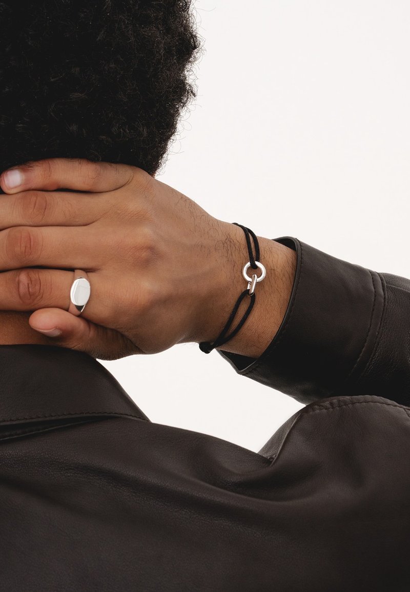 Black cord bracelet featuring a silver circular accent, worn on a hand with a silver signet ring. Leather jacket partially visible.