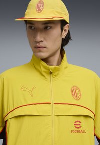 Yellow windbreaker jacket with a high collar, red trim, and Puma logo. Features an embroidered ACM emblem and “Football” text in red.