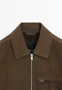 Braune Wildlederjacke mit Kragen, vorderem Reißverschluss und einer Brusttasche. Glatte Textur und minimalistisches Design.