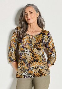 Multicolore blouse met bloemen- en dierenprints, 3/4 mouwen, keyhole-halslijn en zachte stof. Gecombineerd met olijfgroene broek.