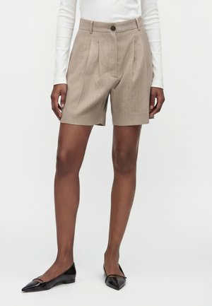 Person iført beige plisserede skræddersyede shorts, en langærmet hvid top og sorte spidse flade sko, står mod en ensfarvet baggrund.