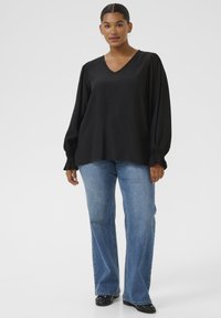 Schwarze Bluse mit langen Ärmeln, V-Ausschnitt und gerafften Manschetten, kombiniert mit hellblauen Schlagjeans und schwarzen Schuhen, vor einem einfarbigen Hintergrund.