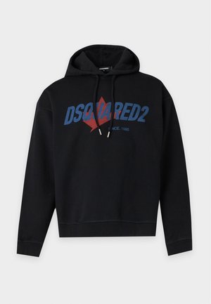 Homme portant un sweat à capuche noir DSquared2, un pantalon blanc et des chaussures noires, debout sur un fond blanc uni.