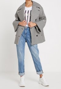 Abrigo a cuadros en blanco y negro con mangas anchas, combinado con una camiseta gráfica blanca y unos vaqueros claros arremangados, completado con zapatillas blancas.