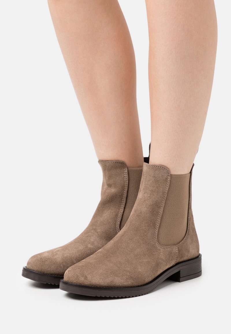 Bottes en daim taupe hauteur cheville avec panneaux latéraux élastiques et semelles noires basses portées sur des jambes nues.