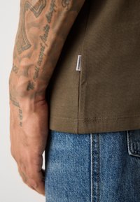 Braț tatuat lângă o cămașă din țesătură ribbită verde cu eticheta Lindbergh, asortată cu blugi denim albaștri.