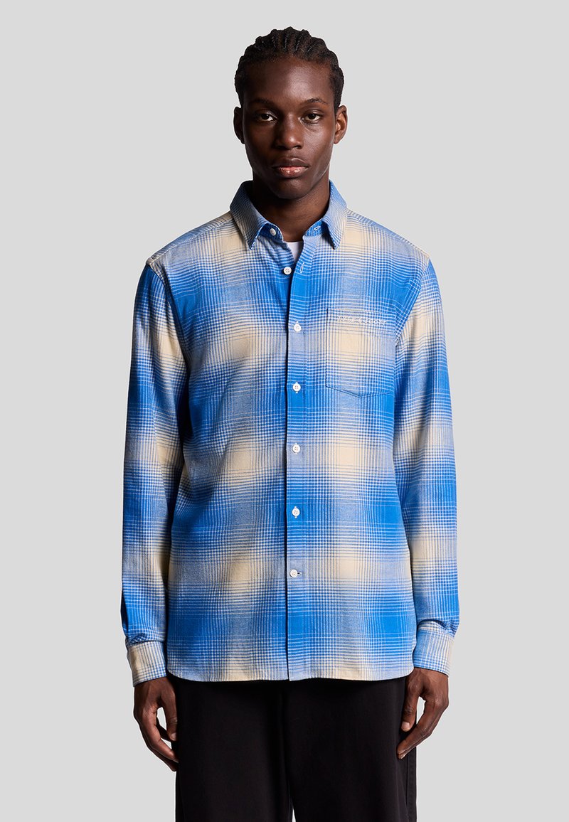 Chemise à carreaux bleue et beige, à boutons, avec un tissu texturé, des manches longues et une poche poitrine, présentant un effet dégradé subtil.