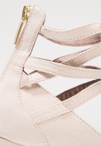Scarpa wedge in suede beige con cinturini incrociati, presenta una zip laterale e una texture liscia. Dettagli di cucitura evidenziano il design.