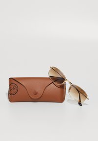 Lunettes de soleil Ray-Ban avec une monture en métal doré et des verres dégradés marron à côté d'un étui en cuir brun texturé avec une fermeture à pression.