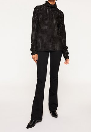 Personne portant un pull à col roulé côtelé noir avec de petits points blancs, un pantalon noir évasé et des chaussures noires pointues.