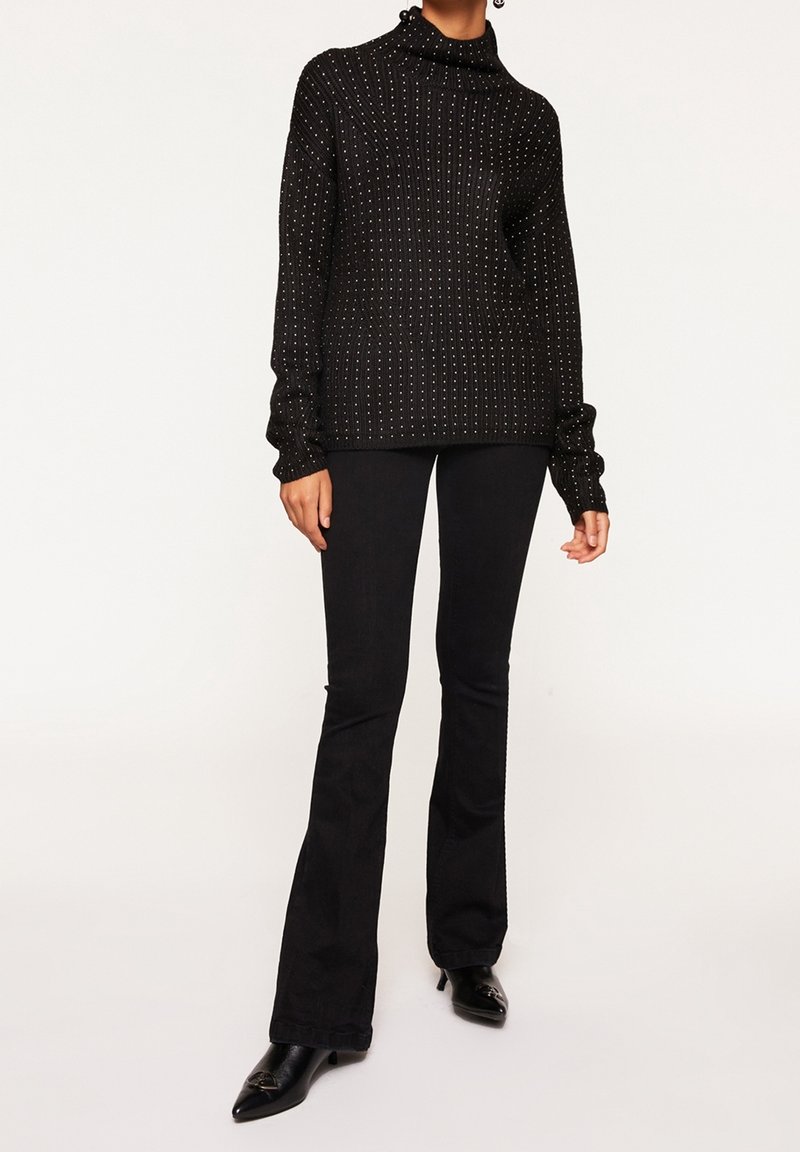 Personne portant un pull à col roulé côtelé noir avec de petits points blancs, un pantalon noir évasé et des chaussures noires pointues.