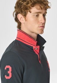 Polo shirt blu navy con colletto rosso a contrasto, dotato di chiusura a bottoni e numero "3" ricamato sulla manica. Tessuto dalla trama liscia.