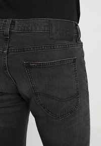 Gros plan d'une personne portant un jean Lee gris foncé montrant la poche arrière et l'étiquette de la marque sur la ceinture.