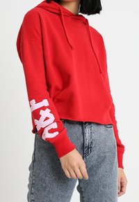 Felpa rossa corta con cappuccio regolabile; presenta lettere bianche sulla manica destra e un tessuto morbido e testurizzato. Abbinata a jeans chiari.
