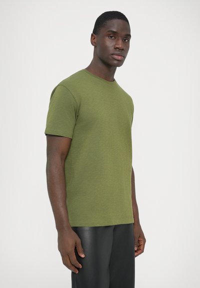 Λαδί κοντομάνικη μπλούζα με στρογγυλή λαιμόκοψη και υφή με ανάγλυφα. Συνδυασμένη με μαύρο λείο παντελόνι. Το μοντέλο στέκεται στραμμένο προς τα αριστερά.