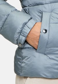 Giacca puffa azzurra chiaro con una texture liscia e lucida. Presenta una tasca laterale e un bottone sui polsini. Design trapuntato con isolamento imbottito.