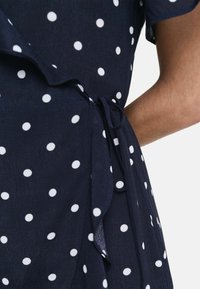 Navyblauwe jurk met witte stippen, wikkelontwerp, gerimpelde randen en een striktaille. Zachte, vloeiende stoftextuur.
