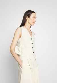 Beige liinapuku v-kaelusega, mustade nööpidega ja küljetaskutega, koos heleda beezist laiade jalgade pükste ja kreemja käteseotiga.