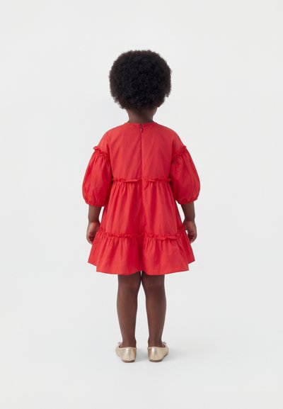 Dítě s krátkými kudrnatými vlasy, oblečené v červených šatech s nabíranými rukávy a zlatých balerínách, stojí zády k fotoaparátu na bílém pozadí.