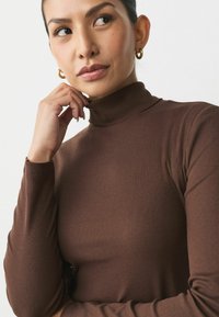 Brown ribbed turtleneck top with long sleeves, featuring a fitted design and a smooth texture, complemented by simple gold hoop earrings.