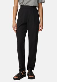Personne portant un pantalon noir taille haute, un haut rayé noir et blanc, et des sandales noires à bout ouvert, debout devant un fond uni.