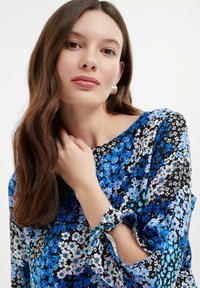 Blusa floreale con base nera e fiori blu, lavanda e bianchi. Vestibilità ampia con maniche legate e texture liscia.
