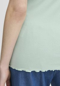 Hele roheline ribadega topp, millel on laineline äär, koos siniste teksapükstega. Suurendatud vaade, mis näitab tekstuuri ja disainidetaile.