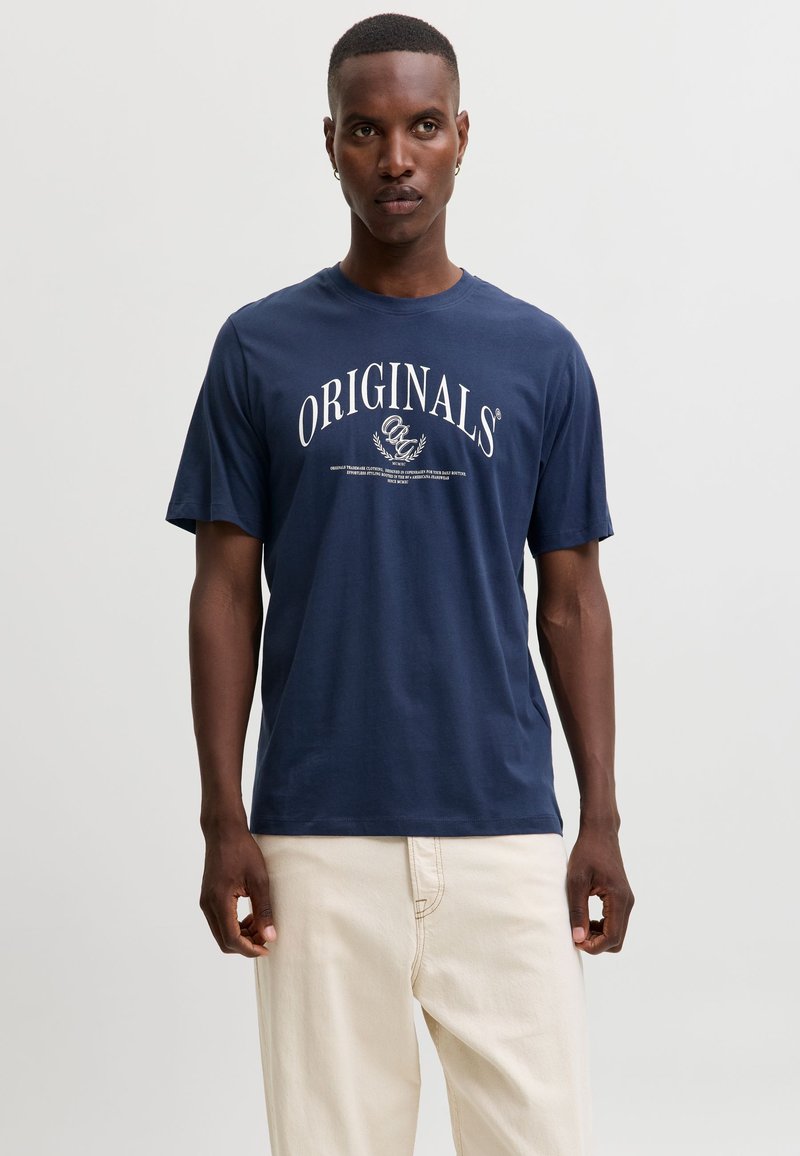 Homme portant un t-shirt bleu marine avec le texte "ORIGINALS" et un pantalon couleur crème, debout devant un fond uni gris clair.