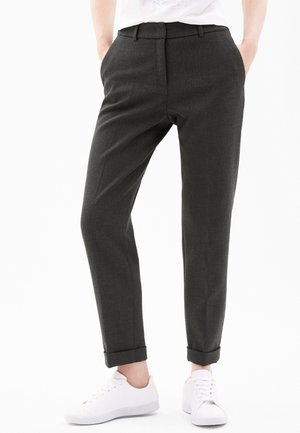 Person wearing dark gray tailored ankle-length pants with cuffs, white sneakers, and a white top, standing with hands in pockets.