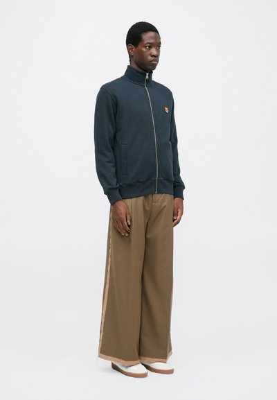 Giacca a zip blu navy con colletto alto, dotata di tasche laterali e un piccolo logo arancione. Abbinata a pantaloni larghi marroni e sneakers bianche.