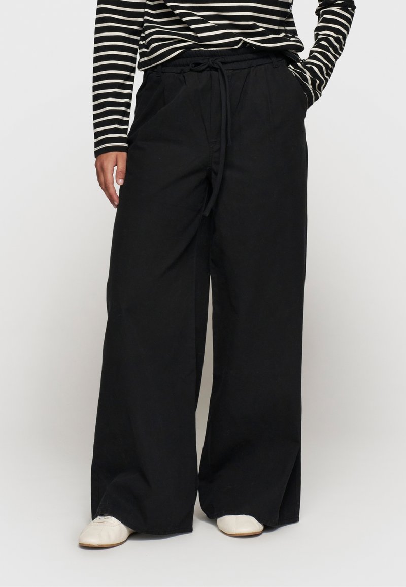 Zwarte wijde pijpenbroek met een koord in de taille, gemaakt van een soepele stof. Gecombineerd met een zwart-wit gestreept shirt met lange mouwen.