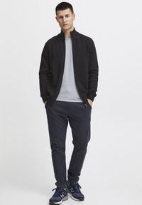 Svart zip-up jacka över en ljusgrå t-shirt, kombinerad med marinblå joggerbyxor och mörka träningsskor. Enkel, avslappnad design.