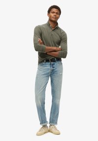Polo de manga larga verde olivo con cuello, combinado con jeans claros desgastados y zapatillas beige, que cuentan con puños enrollados y un cinturón negro.