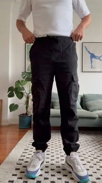 Black cargo pants with multiple pockets, drawstring waist, and tapered leg design. Worn with white and blue sneakers. Indoor setting.