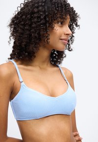 Νέα γυναίκα με σγουρά μαλλιά που φορεί ένα ανοιχτό μπλε υφασμάτινο μπικίνι πάνω μέρος, κοιτάζει προς τα πλάγια με ένα ουδέτερο φόντο.