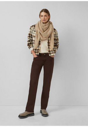 Femme portant une écharpe beige, une veste à carreaux, un haut crème, un pantalon marron foncé et des bottes beiges, debout les mains dans les poches contre un mur gris.