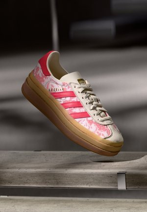Hvid og pink Adidas Gazelle sneaker med gummisål, der svæver over en betonbænk i blødt lys.