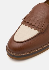 Mocasines de cuero marrón con una punta redondeada de color crema y detalles de flecos. Los acentos cosidos a lo largo de la suela realzan la textura y el diseño.