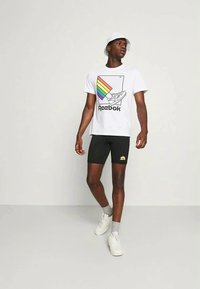 Fehér Reebok póló színes szivárvány grafikával, fekete kerékpáros nadrág, fehér sportcipő és fehér kalap. Egyszerű és sportos kialakítás.