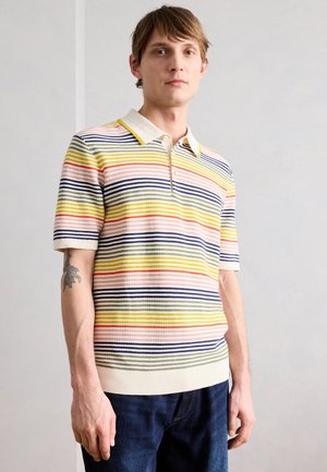 Ung mand med kort hår iført en stribet kortærmet poloshirt og mørke jeans, stående mod en ensfarvet lys baggrund.