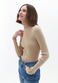 Beige gerippter Rollkragenpullover mit langen Ärmeln, der eine figurbetonte Passform und weiche Textur aufweist, kombiniert mit blauen, hochtaillierten Jeans.