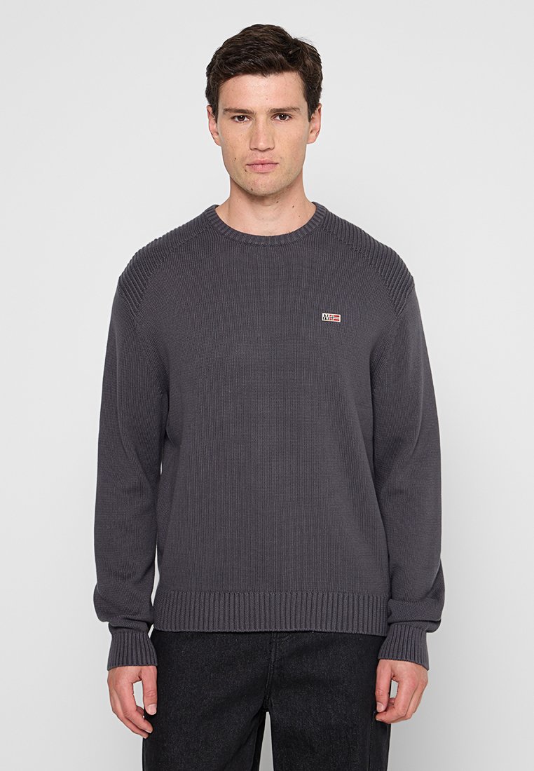 Man in een donkergrijze ribgebreide trui met een klein rechthoekig logo op de linkerkant van de borst, gecombineerd met zwarte broek, staande tegen een witte achtergrond.