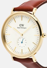 Reloj en tono dorado con una esfera texturada blanca, índices de oro y un segundero azul. Correa de cuero marrón y caja de acero inoxidable.