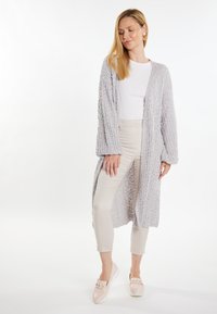 Cardigan lungo grigio chiaro, strutturato, con apertura frontale, indossato sopra una maglietta bianca e pantaloni beige aderenti, abbinato a scarpe slip-on rosate.
