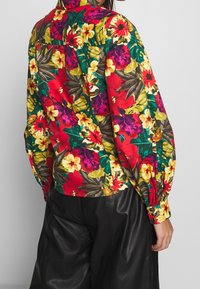 Blouse fleurie aux rouges, jaunes et verts vibrants ; manches longues, col chemise, coupe ample portée avec un pantalon noir.