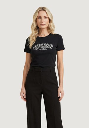 Femme aux longs cheveux blonds portant un t-shirt noir avec le logo "Guess" et un pantalon noir taille haute, debout devant un fond clair uni.