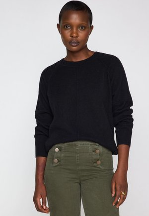 Maglione nero a maglia con maniche raglan, abbinato a pantaloni verde oliva con vita alta e dettagli con bottoni sui fianchi. Texture liscia.