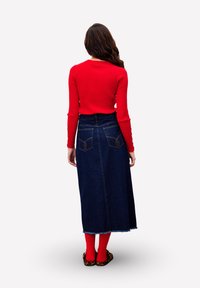 Rotes geripptes Langarmshirt, kombiniert mit einem dunkelblauen Jeans-Maxirock mit Gesäßtaschen und ausgefranstem Saum, ergänzt durch rote Socken und bedruckte Schuhe.