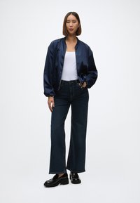 Jūrnieku satīna bomber jackets pār baltu bezdelīgu topu, apvienots ar tumšiem džinsu plaša stila biksēm un melniem platformas loaferiem. Vienkāršs, mūsdienīgs dizains.