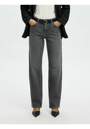 Osoba nosiace čierne rifle s vysokým pásom a opaskom, čierne špicaté topánky, biely top a čierne sako, stojaca s rukami v vreckách.